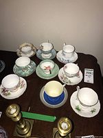 Nine vintage tea cups and matching saucers displayed on a wooden table, showing variety of floral patterns and gold trim details.