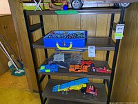 Black shelving unit showing contents: blue two-layer carrying case on upper shelf, assorted metal cars and plastic Smash Up Derby cars on middle shelves, auto transport truck on bottom shelf