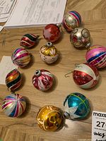 Photo showing thirteen assorted vintage 1960s Christmas ornaments laid out on a wood surface, showing round and teardrop shapes with colorful stripes and glitter details.