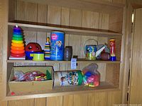 Two wooden shelves built into a wood-paneled wall holding an assortment of vintage toys including a multicolor stacking ring toy, a kaleidoscope, building blocks, toy football, watering can, and LEGO container.