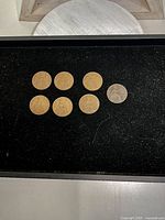 Seven coins displayed on black surface, six bronze and one grey toned