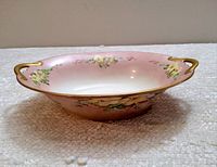 Side angle view showing the oval shape, pink gradient background, floral handpainted designs, and gold handles.