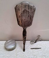Image of grouped vintage vanity items including silver powder compact, hand mirror, eyelash curler, and eyebrow plucker.
