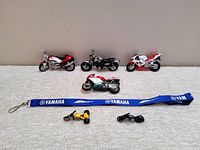 Seven diecast and plastic toy motorcycles arranged in two rows with a blue Yamaha lanyard in front on white fabric background.