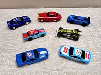 Seven assorted toy slot cars arranged on a white surface showing styles and colors.