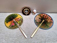 Two reversible Asian-style hand fans with colorful nature-themed designs, one predominantly green and yellow, the other orange and gold, alongside a small decorative egg-shaped milk glass perfume bottle and a round 24kt gold Chokin pin dish.