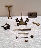 Photo showing a hammer-shaped bottle opener, small brass abacus, decorative clip with animals, and assorted small brass screwdrivers and fittings against a white cloth background.