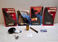 Four vintage splash guards still in original packaging, brands visible Dodge (2), Oldsmobile, and Ford, displayed upright side-by-side with other items such as belt and key ring holder, patch, and repair kit.