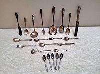 Vintage silverware assortment laid out horizontally. Includes spoons, forks, small butter knife, ladles with visible tarnish and patina.