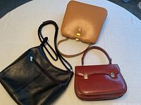 Three leather handbags: modern black bag, vintage tan and reddish brown bags laid on white surface.