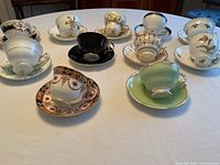 Front view of 11 assorted bone china teacups with matching saucers arranged on a white tablecloth. Mix of floral designs and colors including black, green, white, and multi-color florals.