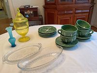 Overall view of yellow candy jar, blue vase, clear glass platters, and green ceramic mugs, saucers, and plates on white tablecloth