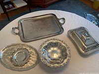 Photo showing four assorted silver plated serving dishes on a table: rectangular tray with handles, oval tray, ornate bowl, and covered dish.