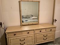 Front view of cream-colored dresser and attached rectangular mirror reflecting a bed.