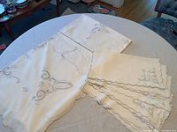 Table displaying the rectangular white linen tablecloth with detailed gray embroidery and scalloped edges, along with the 8 matching napkins stacked nearby, illustrating the condition and appearance of the set.