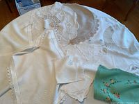Several table covers laid out showing various embroidery, lace, and floral designs including a green embroidered cloth.