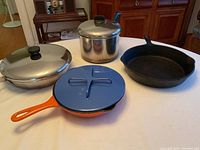 Lot of cooking pots and pans on white table: includes orange Le Creuset skillet with lid, stainless steel steamer pot with lid, large cast iron pan.