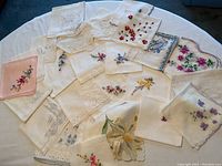 Full group of eighteen vintage embroidered handkerchiefs laid out on a round white table showing various floral embroidery patterns in different colors.