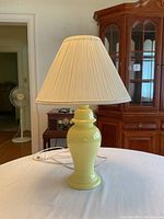Yellow ceramic table lamp with an ivory pleated shade on top of a white table, lamp is lit in one image showing the shade condition.