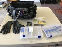 Full view of the fishing carryall with pliers in one of the external pockets, heavy-duty gloves, and two clear plastic tackle boxes on a table.