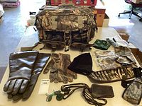 Overall view of hunting supplies including Avery camo carrying case, various gloves, face and neck warmers, shell belt, and whistle.