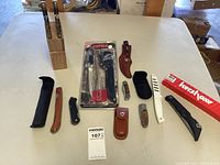 Overview of all knives and sheaths on table including Rapala Soft Grip Fillet Knife in packaging and multiple folding knives and sheaths