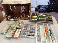 Full lot overview of fishing lures including boxed Flatfish lures, plastic organizers with multiple small lures, and loose metal jigging rappers in cardboard box.