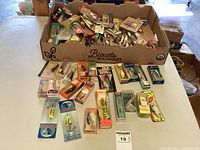 Wide view of box with many opened and unopened fishing lures, some arranged on table.