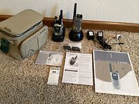 Photo showing two Motorola Talkabout two-way radios model T7100 and MC225R in camo pattern seated in charging docks with accessories including power adapters, antennas, user manuals and a soft carrying case.