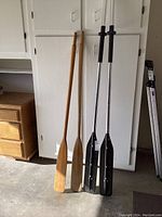 Full view of two wooden paddles and two aluminum paddles leaning against a white cabinet in a room.