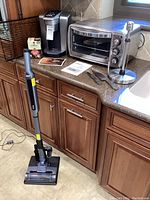 Lot of small household appliances and kitchen items on countertop and floor, including Keurig K-Elite coffee maker, Oster toaster oven, Shark floor sweeper, paper towel holder, and cutlery.