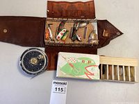 Photo showing Pflueger fly fishing reel, a brown leather lure box open to show 12 assorted lures, and a Mepps cardboard box with 5 lures.