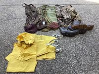 Image showing all the items in the lot laid out: 4 pairs of waders in different camouflage patterns plus solid olive with reddish boots, a yellow rain jacket spread out, and multiple pairs of boots visible.