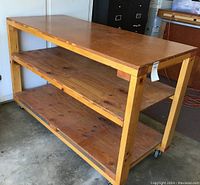 Full side view of wooden workbench showing top surface and two shelves beneath, all constructed of natural wood with visible grain and knots, supported by a simple frame on casters.