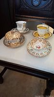 Four pairs of vintage china teacups and saucers arranged on a chair seat, showcasing different floral and gold accent designs.