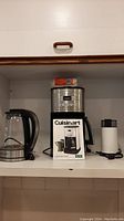 Photo showing all three appliances: Cuisinart coffee maker with packaging, Hamilton Beach electric kettle, and Braun coffee grinder arranged on a shelf.