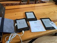 Photo showing two Kindle e-readers and one Kobo e-reader with Kobo owner's manual on wooden table.
