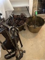 Photo displaying wrought iron fireplace tool set, brass bucket filled with pine cones, and plain brass bucket with a poker inside, all on carpeted floor.
