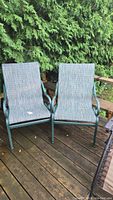 Two metal and mesh patio deck chairs on wooden deck with green foliage background