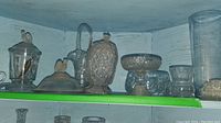 Shelf view of assorted pressed glass items including vases, candy dishes, and flower frog.