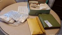 Table showing green hardcover album, cardboard box with envelopes of stamps, clear plastic bags of stock sheets