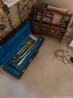Full view of trumpet in blue-lined hard case with harmonica on case floor, wooden recorder leaning on sheet music rack with multiple sheets.