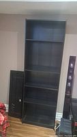 Tall black book shelf unit with several shelves visible, pictured against a wall near a black cabinet and other furniture.