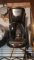 Black & Decker coffee machine with glass carafe on wire shelf, showing front and top of unit with control buttons and logo.