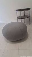 Grey pouf ottoman photographed against a white wall and tile floor, showing its full rounded shape and stitched panel design.