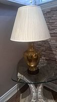 Full view of the Frederick Cooper brass lamp with a cream pleated lampshade on a glass side table in a corner near a stone-textured wall.