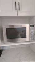 Stainless steel Panasonic microwave oven closed on kitchen countertop beneath white cabinets.