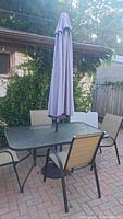 Outdoor rectangular glass table with black metal frame and light purple umbrella centered through table hole, surrounded by outdoor chairs with beige sling fabric.