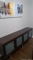 Photo showing the entire front and top surface of the rectangular wooden sideboard with frosted glass doors. Sideboard is on a wood floor against a wall with a painting above.