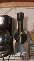 Photo of a black and silver sorbet maker next to a coffee maker on a wire shelf. The sorbet maker has a vertical feeding chute with a green button and dispensing spout.